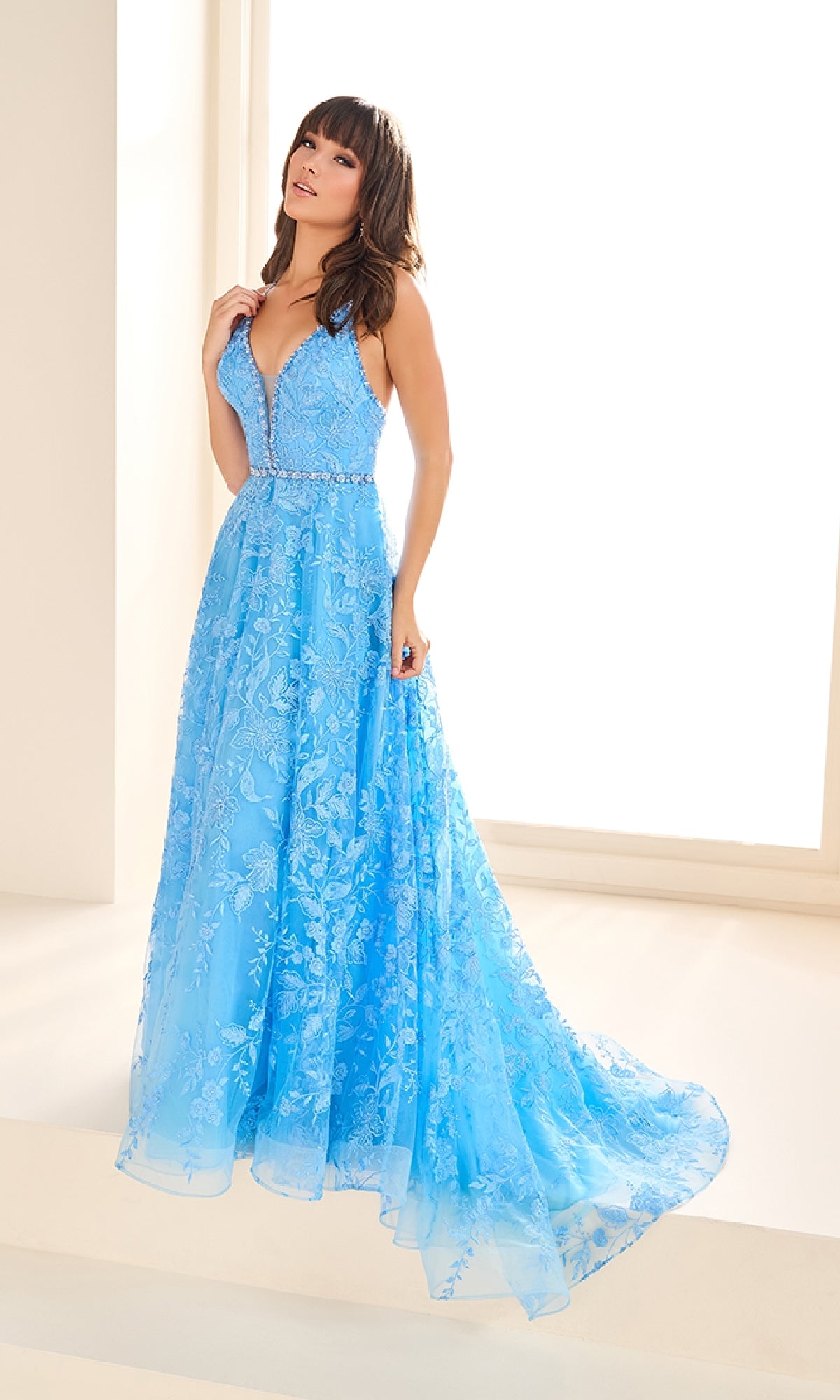 Indoors, a woman models the Ellie Wilde EW36088 long prom dress in light blue embroidered tulle, featuring lace details, a beaded waistband, fitted bodice with thin straps, A-line floor-length skirt with pockets.